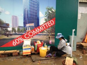 A vendor selling drinks from a bucket, sitting on a board, surrounded by a puddle of mud, with a sign advertising a new highrise development behind her.